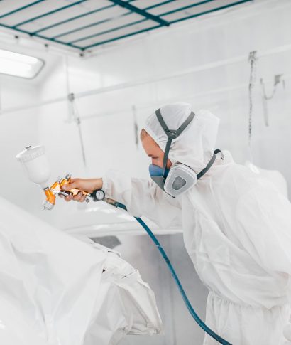 Painter in protective uniform painting car body elements in paint chamber.