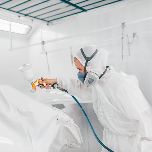 Painter in protective uniform painting car body elements in paint chamber.